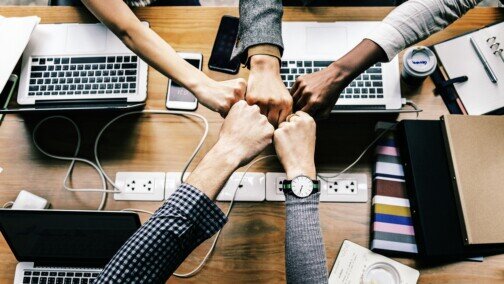 Five people doing a fist bump.  Open laptops, cellphones, and coffee